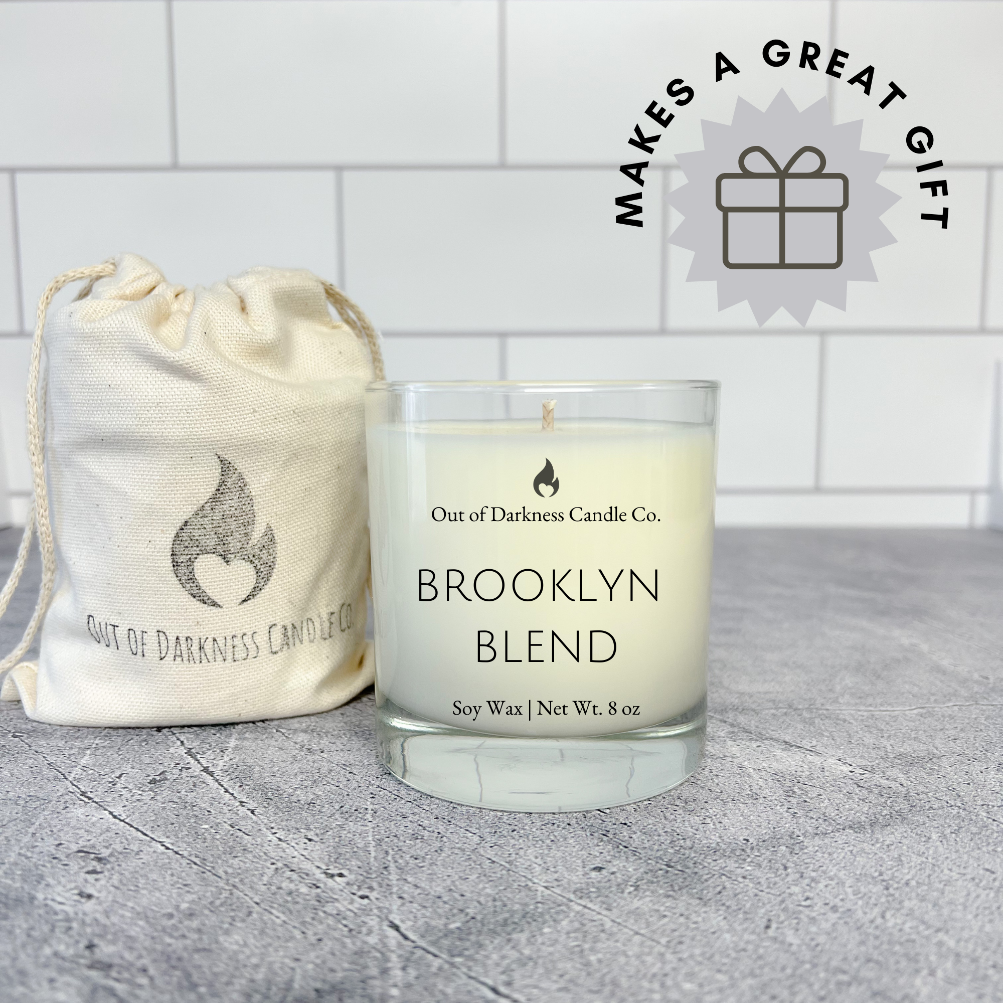 white tile background with present graphic in the corner that says makes a great gift - photo of a glass jar candle that says Brooklyn Blend in all caps with a wrapped candle next to it - wrapped in a muslin bag that has the company logo a black flame and the company name Out of Darkness Candle Co