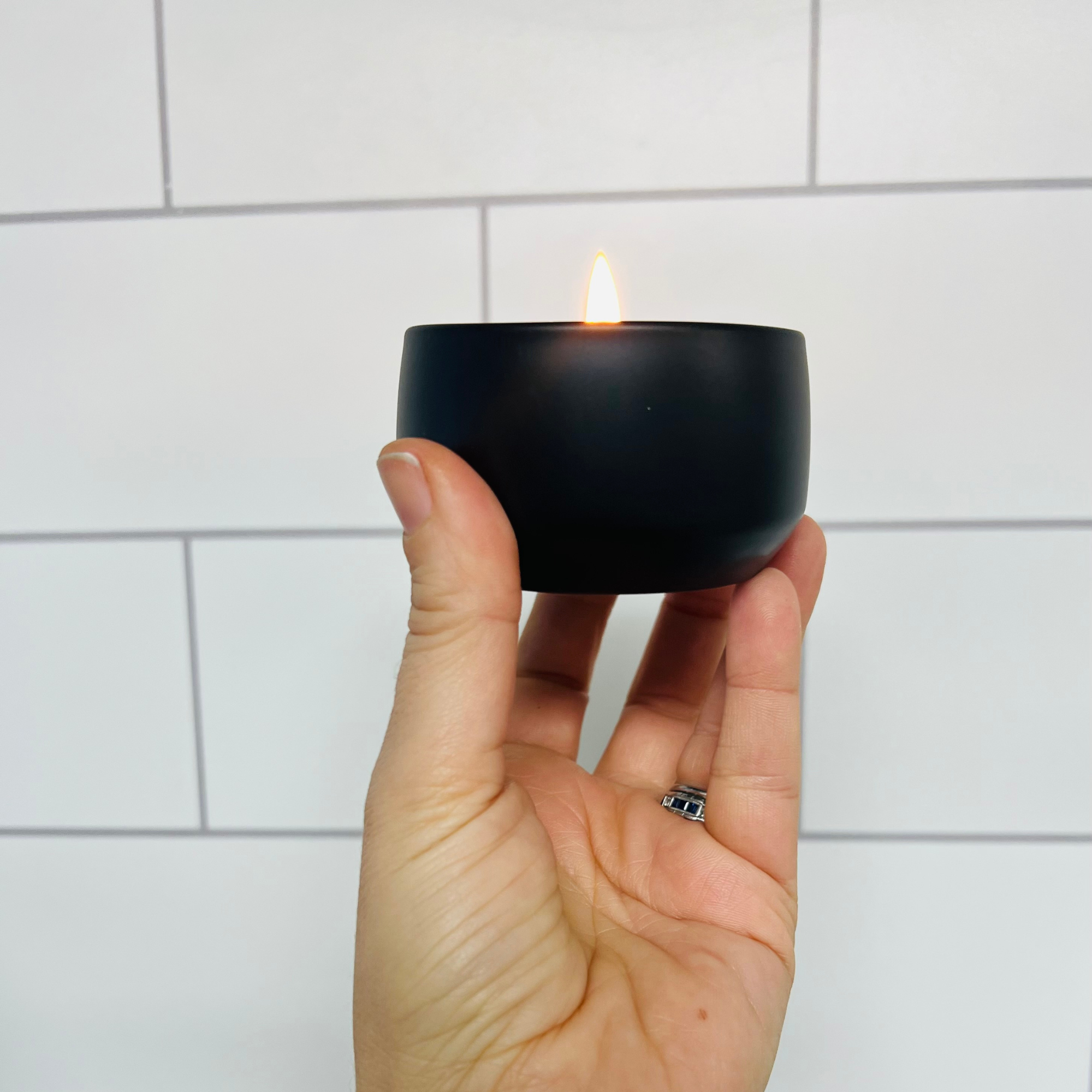 White tile background with a hand holding up a small black matte tin candle that is lit