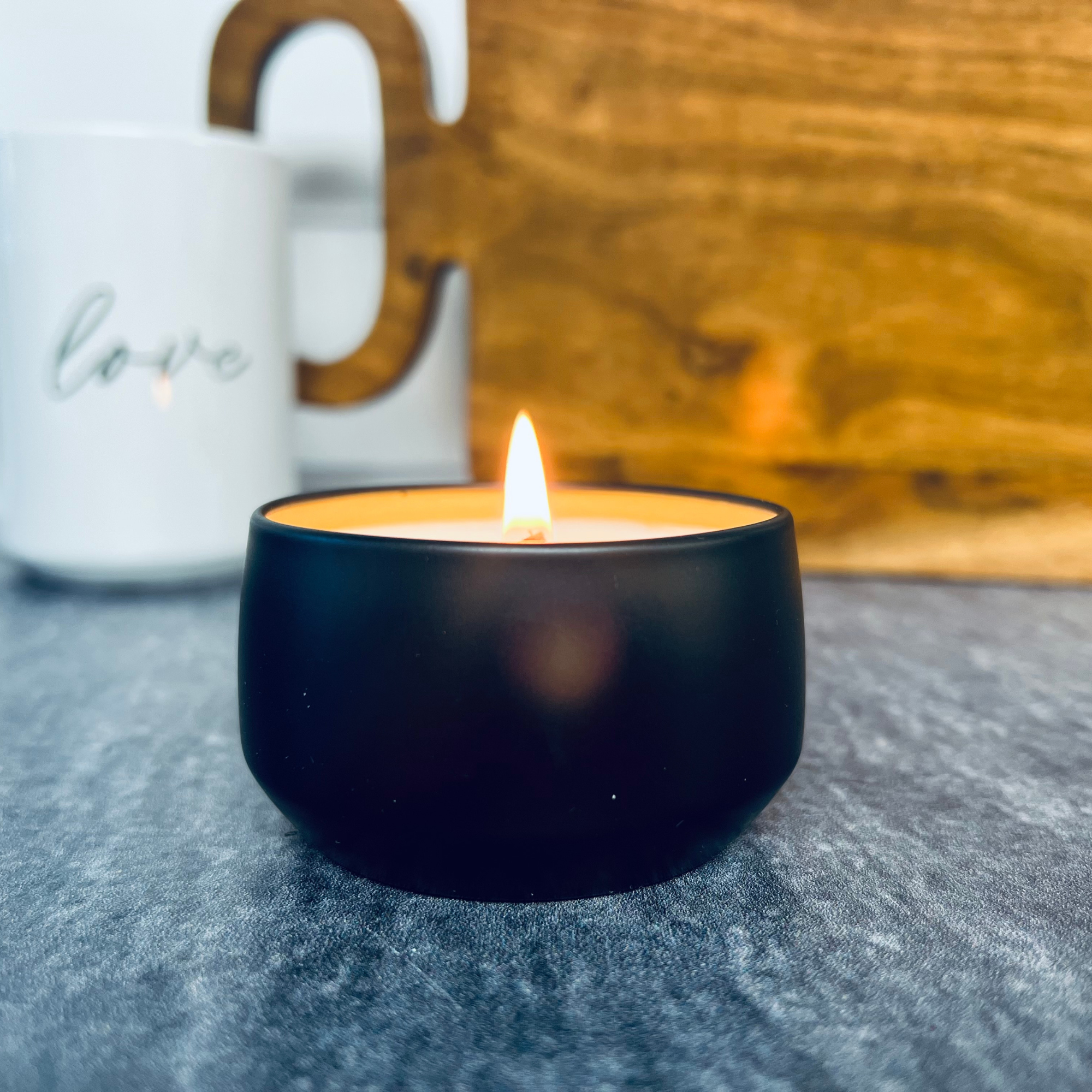 Black Matte Small Candle Tin Lit on a gray table top with a wooden cutting board and a white mug that says love in the background