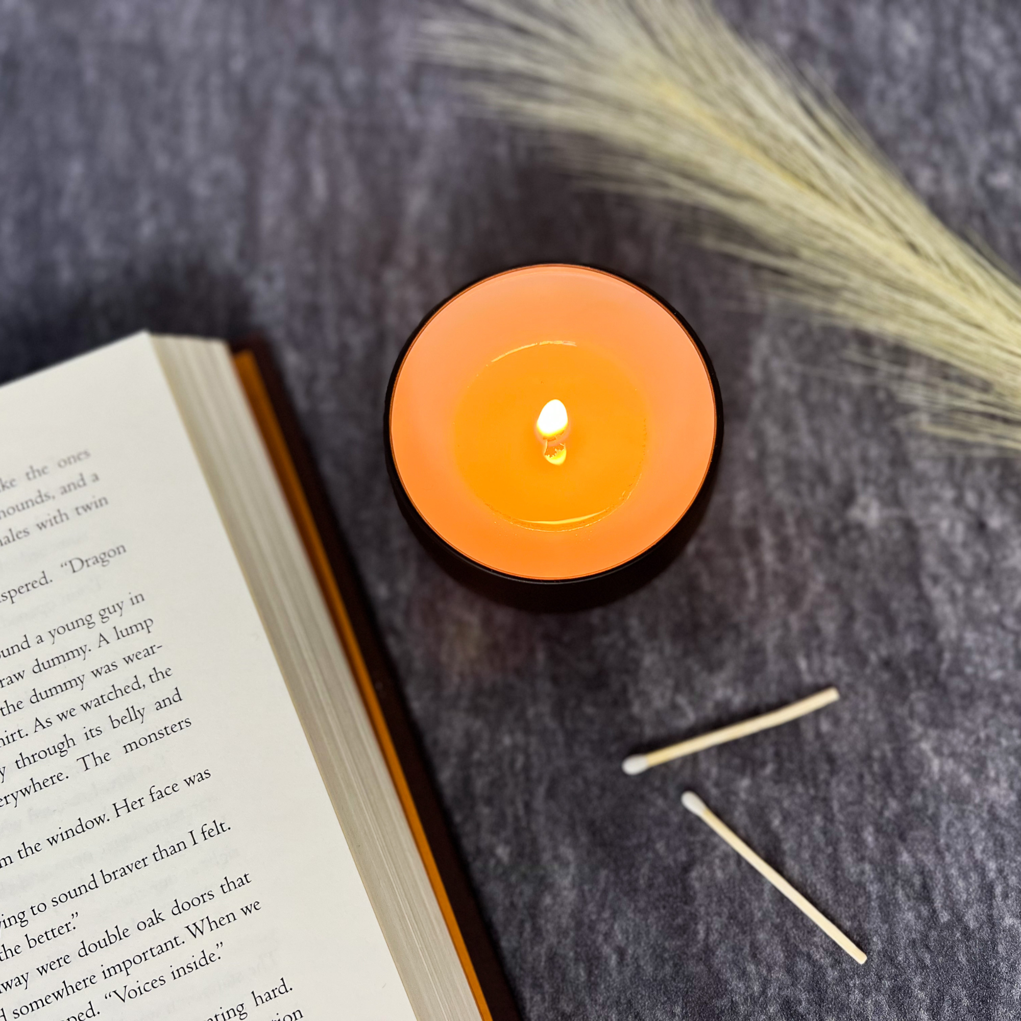 A small candle lit surrounded by two wooden matches a feather and an open book on a gray table