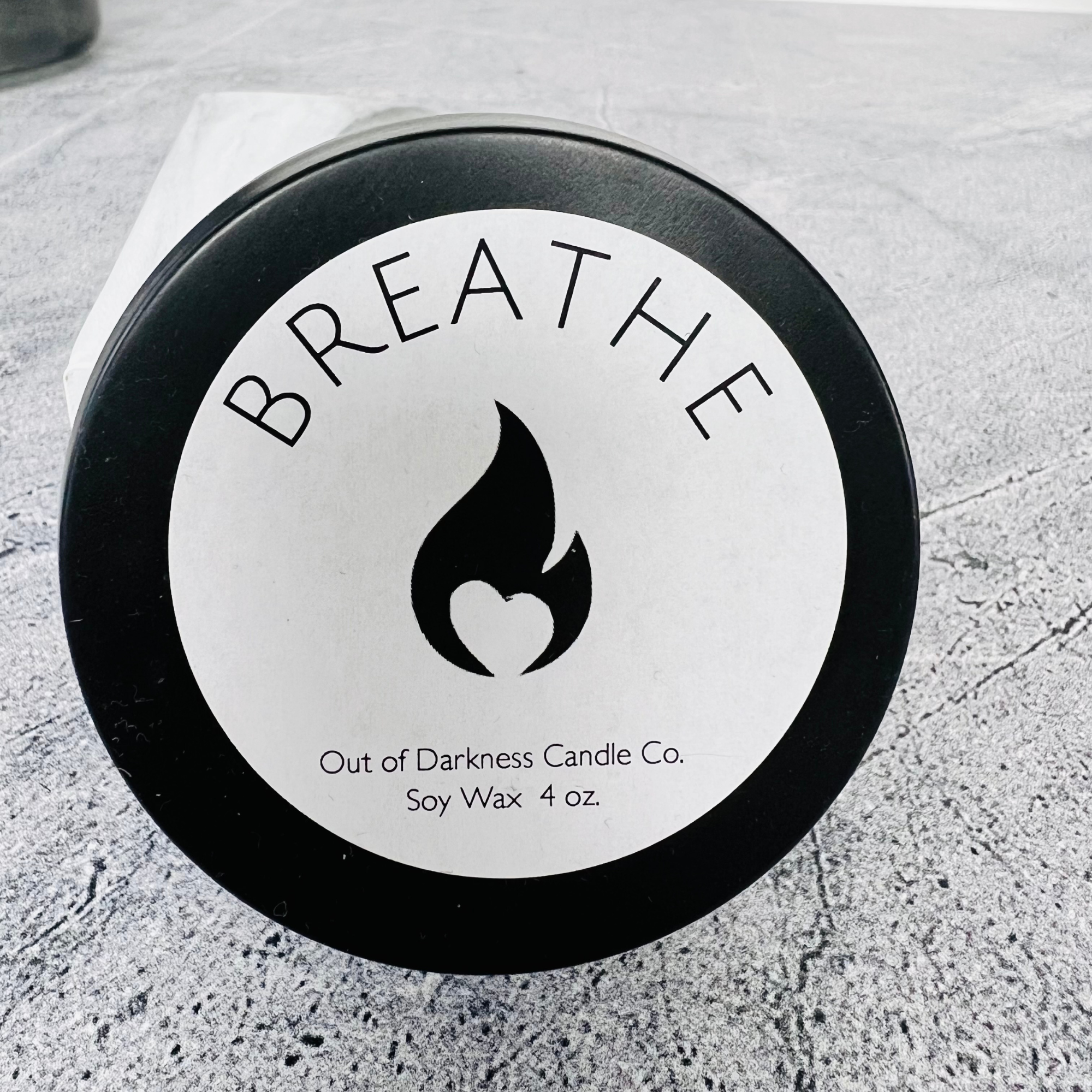 Close up of a Black Matte Candle Tin with a white label on the lid that has a Black Flame - company logo in the middle and says Breathe in all caps around the flame on the bottom of the label it states Out of Darkness Candle Co. Soy Wax 4 oz