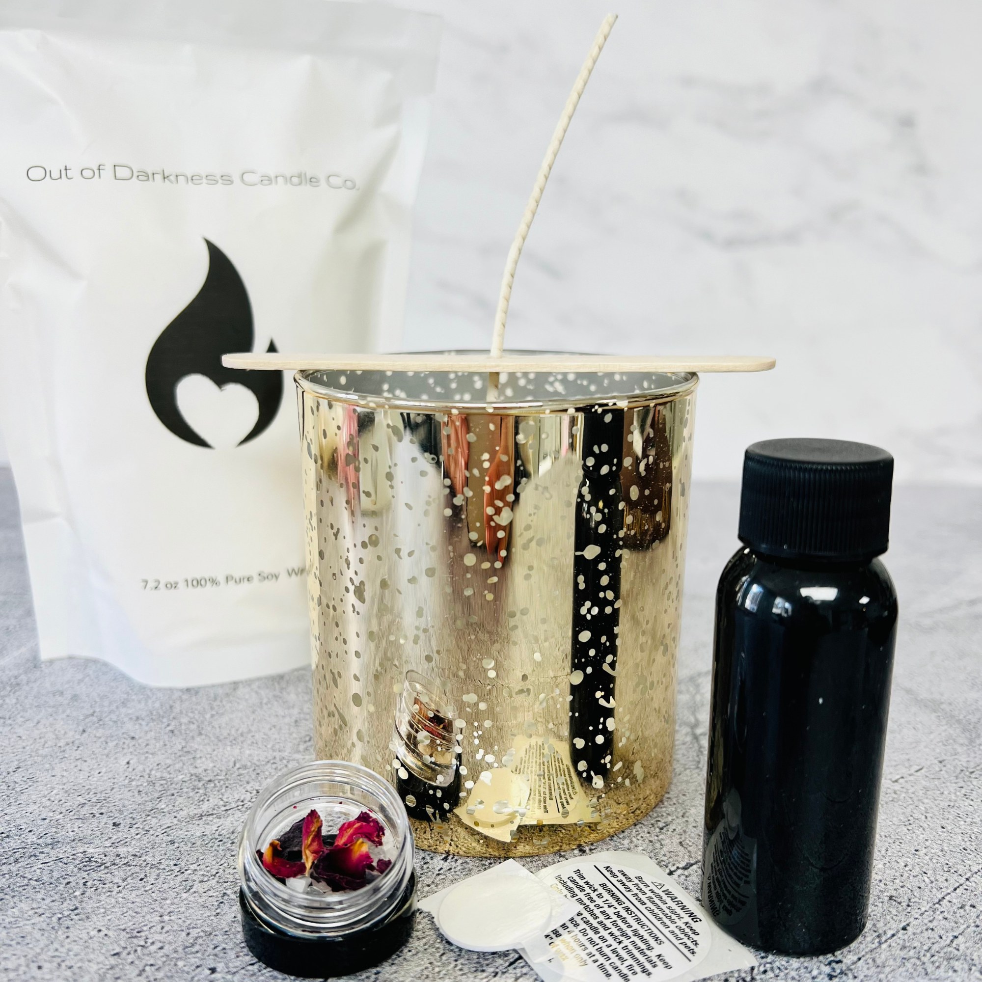 Gold fleck vessel with a cotton with and wooden holder surrounded by a black bottle, a warning sticker, a small container with rose petals and a white bag with black flame company logo on it