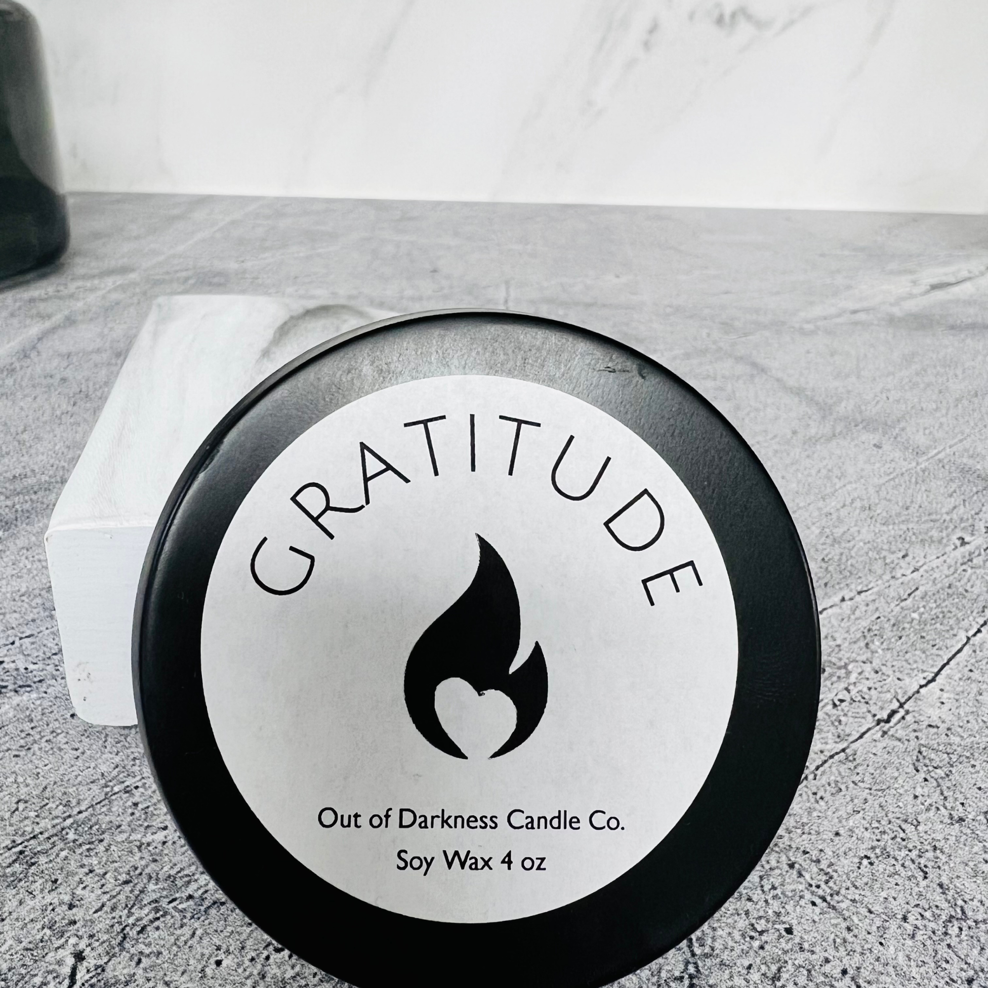 Black Candle Tin with White label on top black flame logo in the middle says gratitude around the flame , bottom of label says Out of Darkness Candle Co. Soy Wax 4 oz 