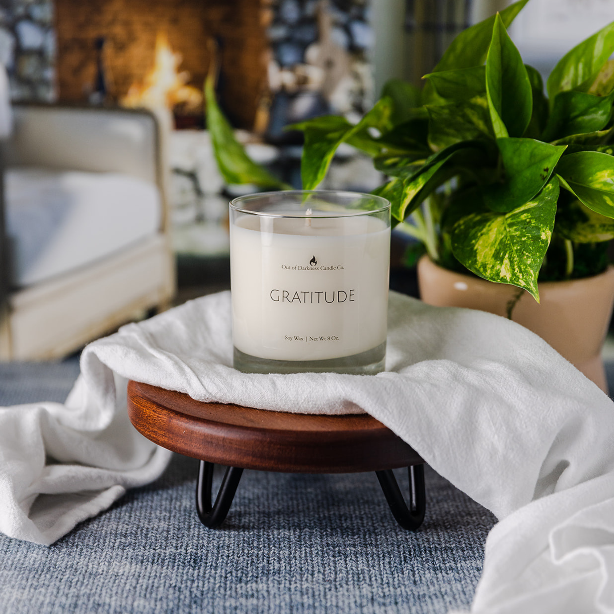 Glass jar candle says gratitude in all caps on a wooden riser with a white scarf draped underneath it, candle is placed in a room with a green plant a white cozy chair and a fireplace with a fire going 