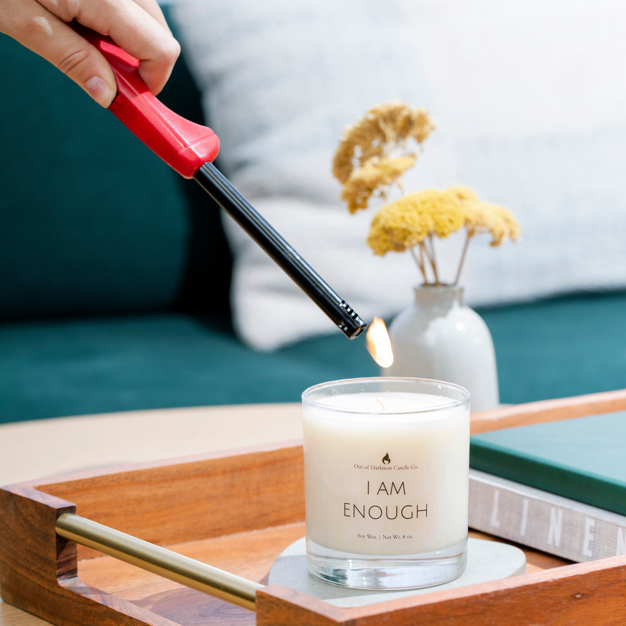 clear glass jar candle in a living room setting on a wooden tray with book and a flower behind it being lit with a lighter