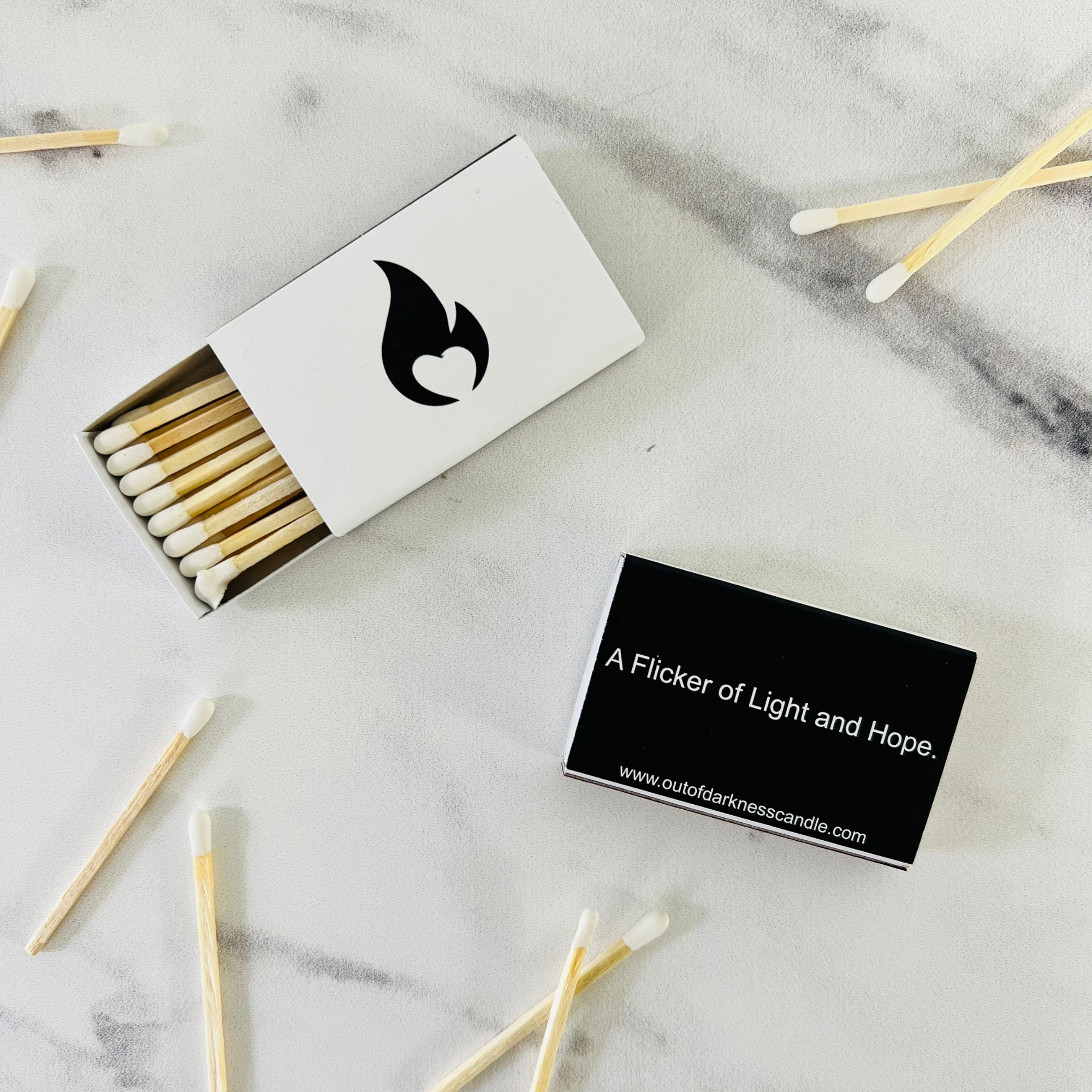 Two boxes of wooden matches on a marble table top with wooden matches around them- One box is white with a black flame and the other matchbox is black with white text reading A Flicker of Light and Hope  