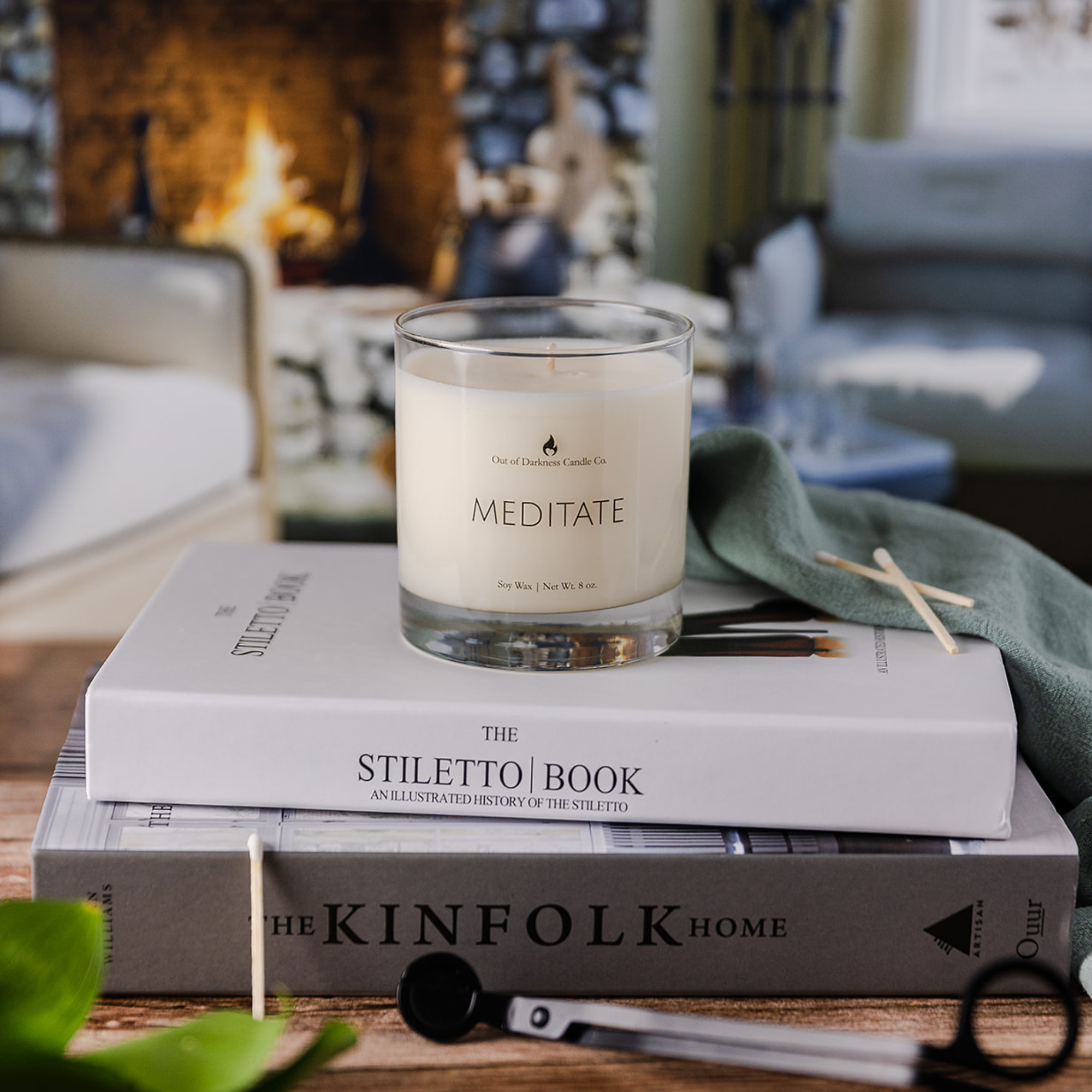 Glass Jar Candle says meditate on top of two hard covered books with a wick trimmer in front and a living room with a stone fireplace and fire in the background