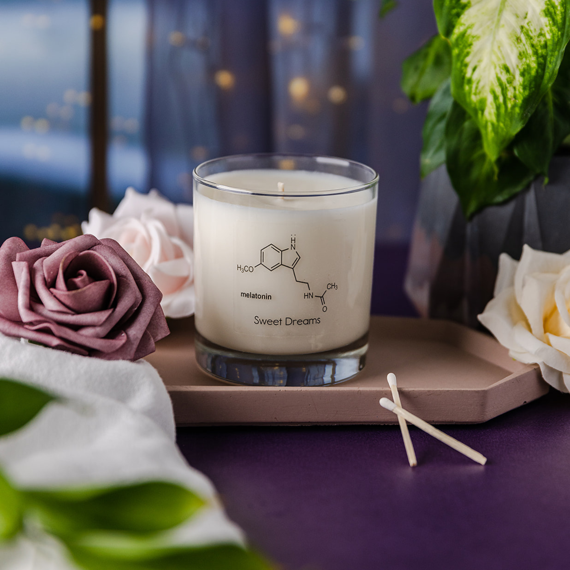 Glass Jar Candle with the molecule melatonin says sweet dreams. It is surrounded by beautiful roses on a dark purple table, two wooden matches in front of the candle and a curtain with fairy lights in the background
