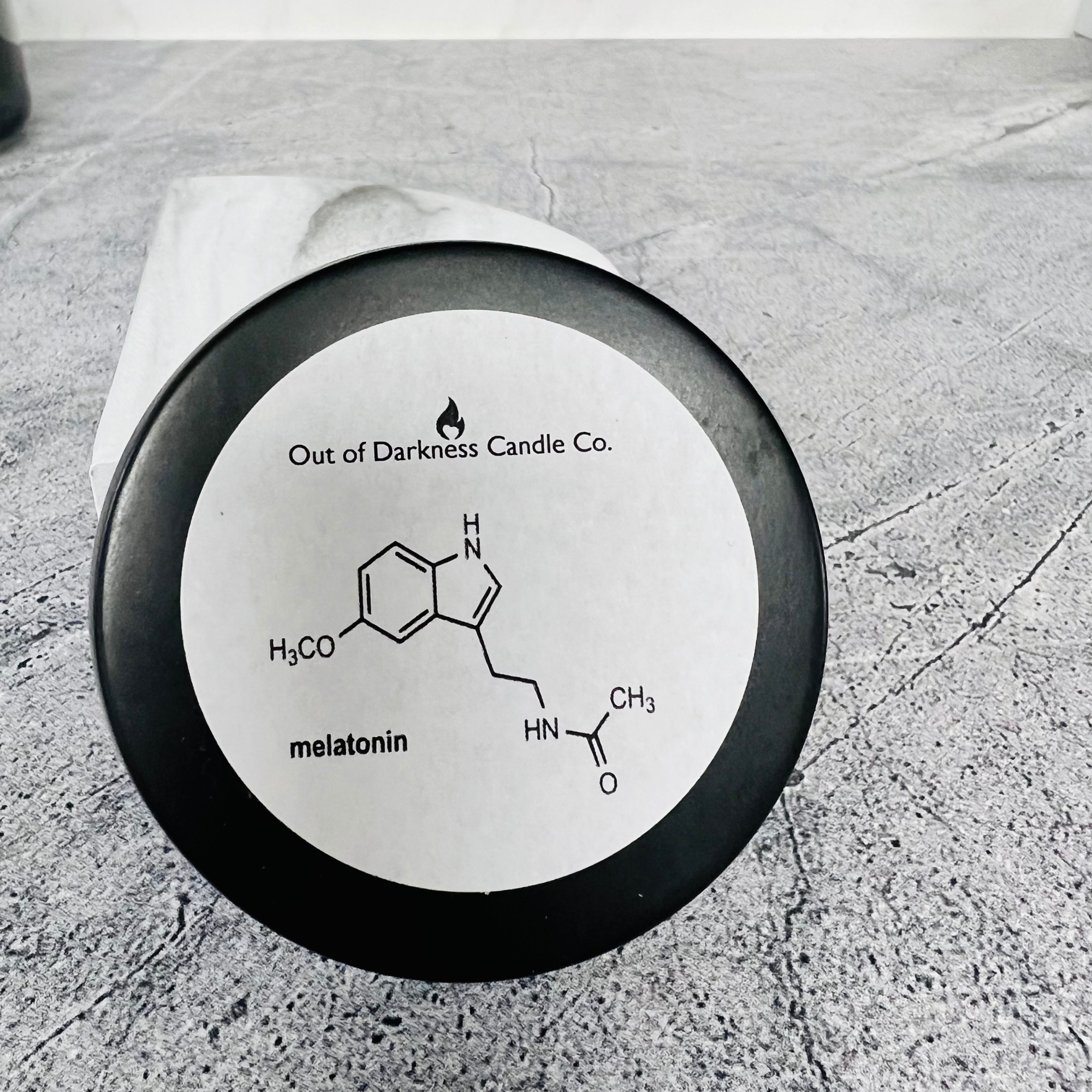 Black Candle Tin with a white label that shows the molecule for melatonin -says melatonin on a gray tabletop