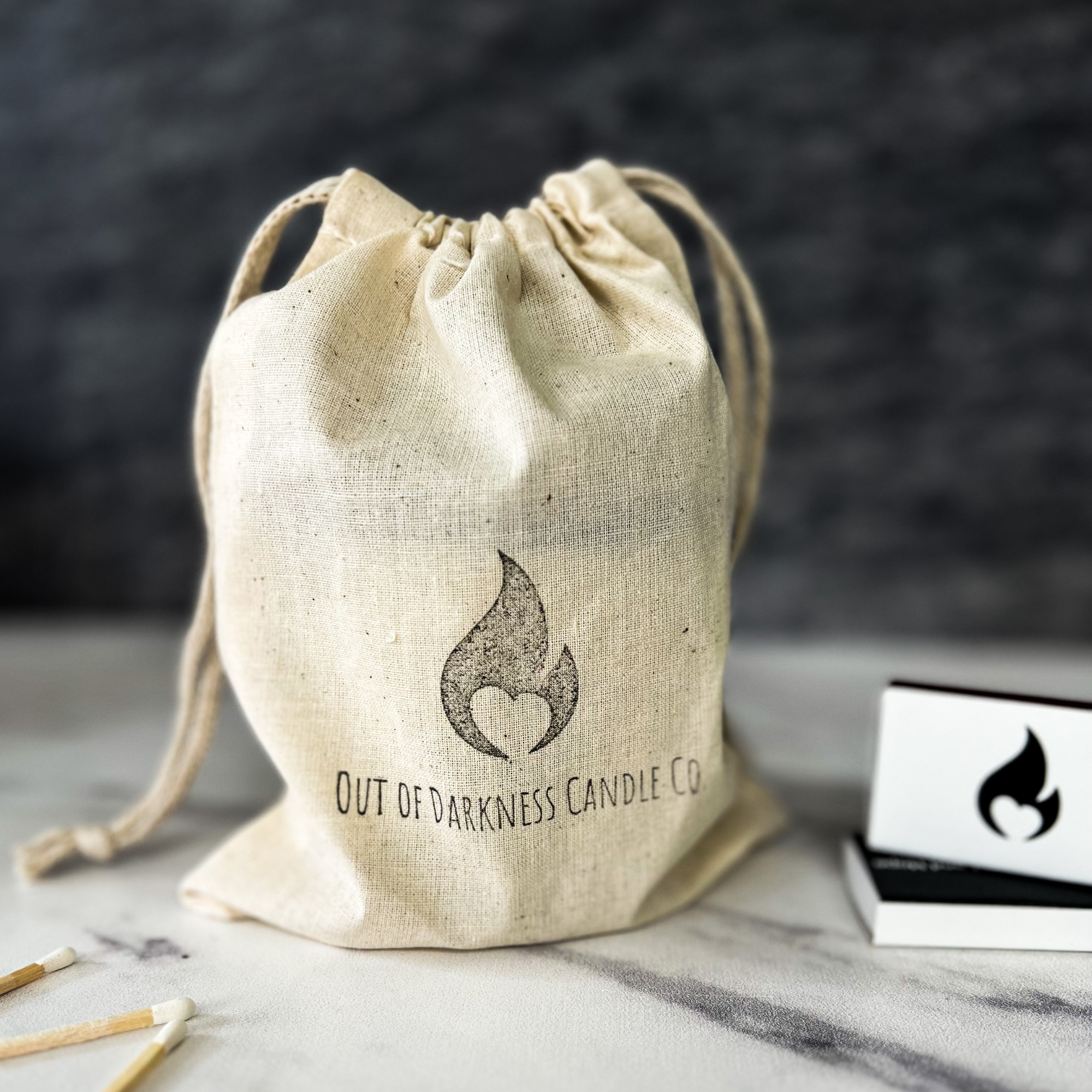 Off White Muslin Bag that says Out of Darkness Candle Co and has the company flame logo on top of that- next to the bag are a book of wooden matches