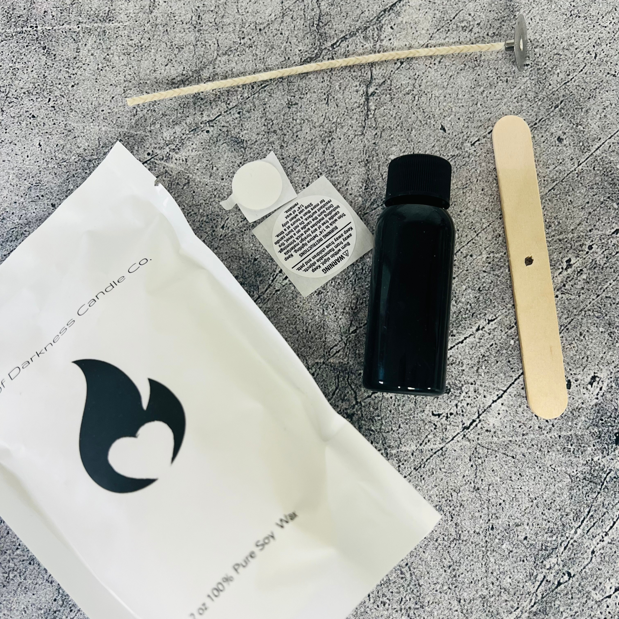 Cotton Wick, Wooden Wick Holder, Warning Label, Wick Sticker, Black Bottle with top and White Bag with Black Flame Logo on front laid out on a gray table