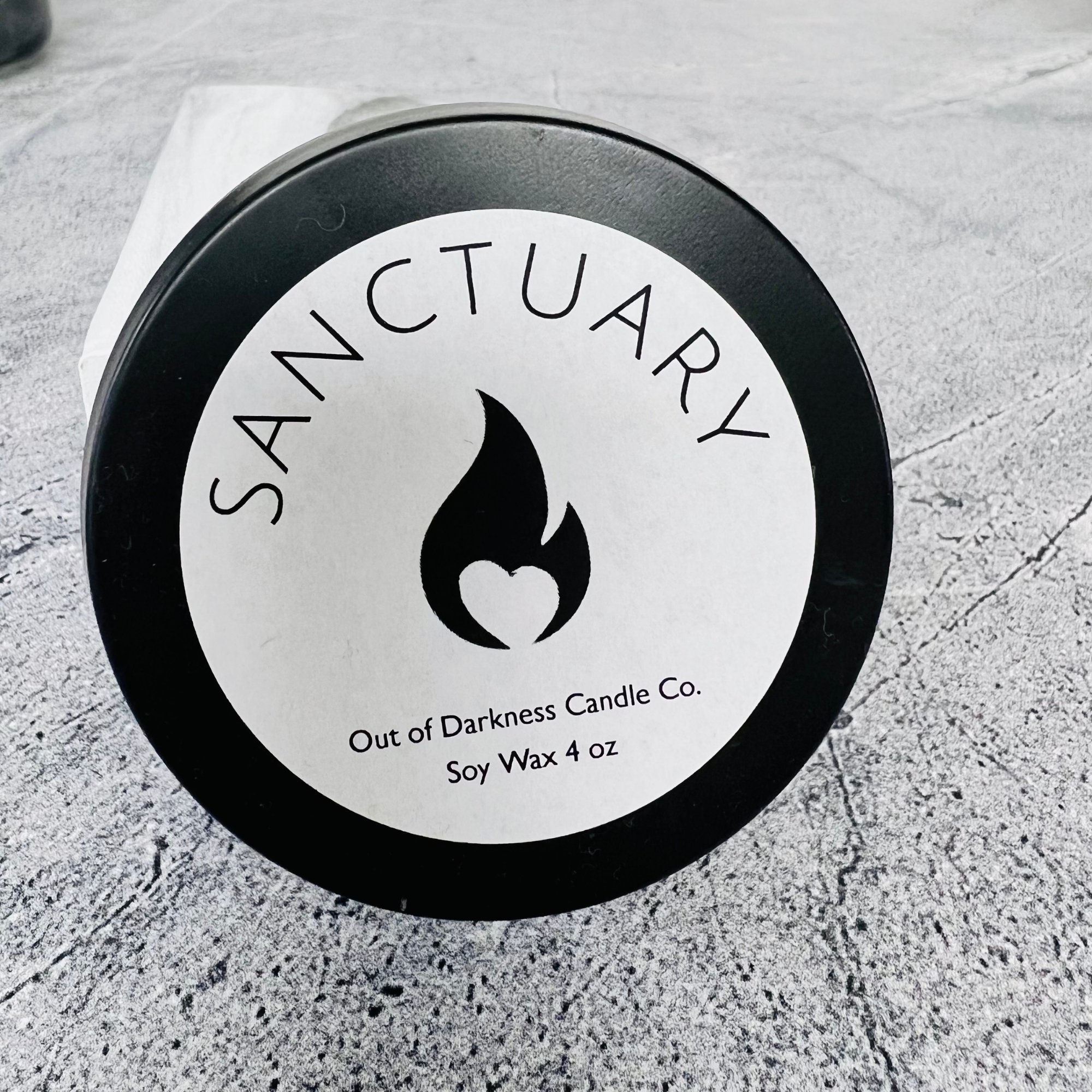 black candle tin with white label - black flame logo in the middle says sanctuary above the flame and below the flame says out of darkness candle co soy wax 4 oz
