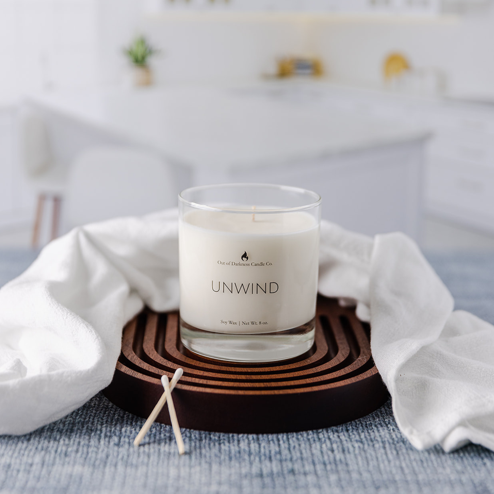 clear glass jar candle says unwind on a wooden riser draped by a white scarf two wooden matches on a blue tablecloth kitchen in the background