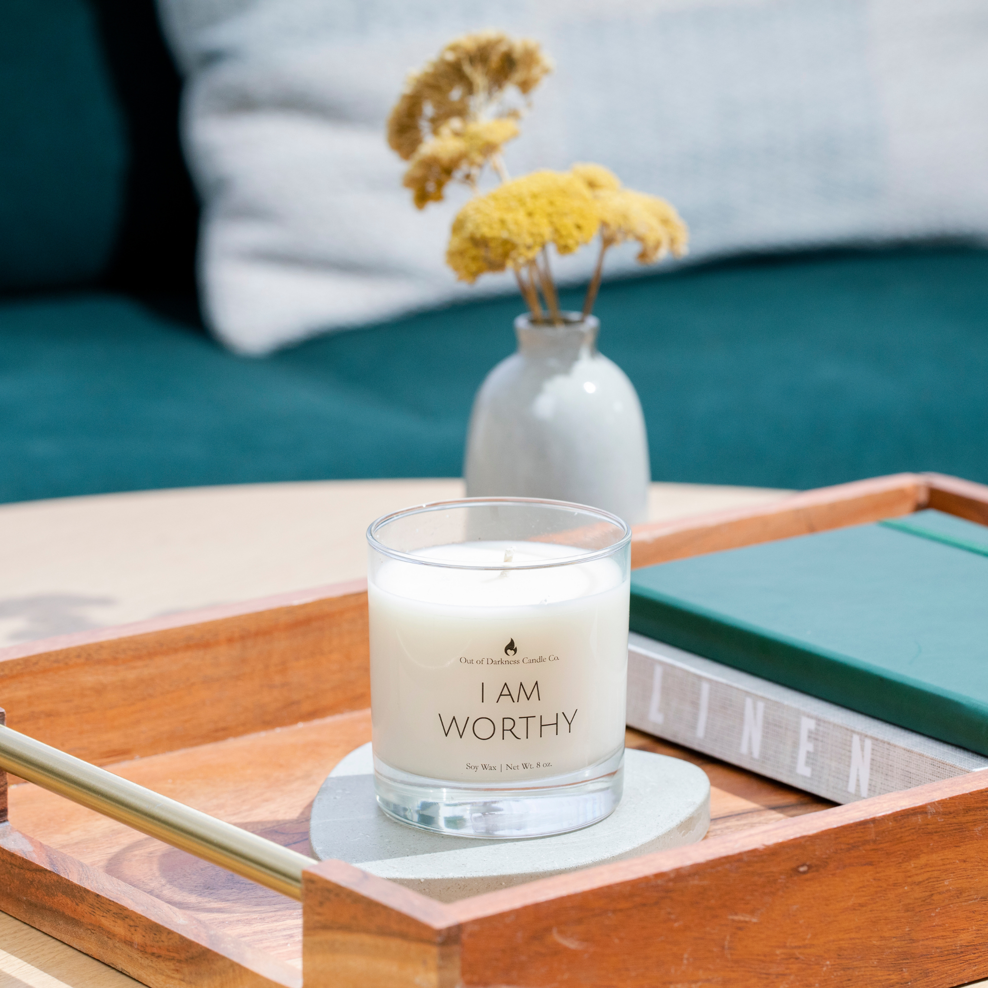 clear glass jar candle says I am worthy in a living room setting on a wooden tray with books and a yellow flower